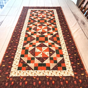 May include: A rectangular quilted table runner featuring a central geometric pattern of orange, dark green, and cream squares and triangles. The border displays a dark brown fabric with a repeating print of small houses and autumn leaves.