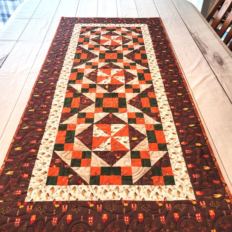 May include: A rectangular quilted table runner featuring a central geometric pattern of orange, dark green, and cream squares and triangles. The border displays a dark brown fabric with a repeating print of small houses and autumn leaves.