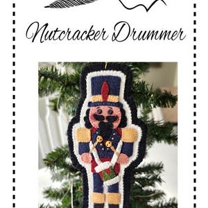 Könnte beinhalten: Ein Filz-Nussknacker-Trommler-Anhänger mit einer rot-weiß-blauen Uniform, der Schlagstöcke hält. Der Anhänger hängt an einem grünen Weihnachtsbaum.