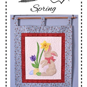 Può includere: Un arazzo da parete trapuntato blu e rosso con un coniglio, un narciso e uova di Pasqua. L'arazzo ha un bordo inferiore a forma di conchiglia e il testo "Spindles Designs by Mary and Mags Spring" in alto.