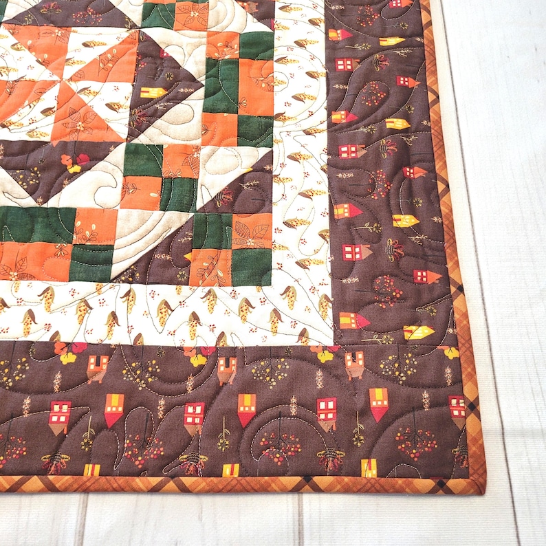 May include: A quilted fabric piece featuring a patchwork design with orange, dark green, and cream squares. The border is a dark brown fabric printed with small houses and trees, finished with a plaid binding in shades of orange and brown.