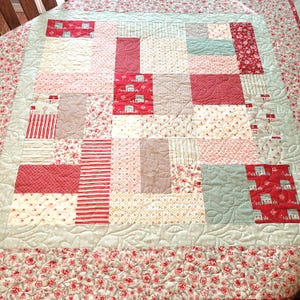 Peut inclure: Une courtepointe patchwork avec divers tissus rouges, cr&egrave;me et bleu clair. Elle pr&eacute;sente des carr&eacute;s et des rectangles avec des motifs floraux, ray&eacute;s et de maisons. La bordure est un imprim&eacute; floral rouge et cr&egrave;me.