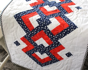 May include: A white quilted table runner with a red, white, and blue geometric pattern. The pattern features stars and squares. The runner has a black and white striped border.