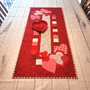 May include: A red quilted table runner with a heart-themed design. The runner features a central column with various red and white patterned squares, heart appliqués, and a red heart-shaped bowl. The edges are trimmed with a white and red striped border.