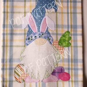 May include: A white tea towel with a blue and yellow plaid pattern. A patchwork gnome with bunny ears is appliqued on the towel. The gnome is holding a green heart and surrounded by colorful eggs.