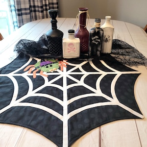 May include: A black fabric table runner featuring a white spiderweb pattern and a green spider applique wearing a purple hat. Behind the runner are five decorative glass bottles in black, white, and maroon, some with spider and bat motifs.