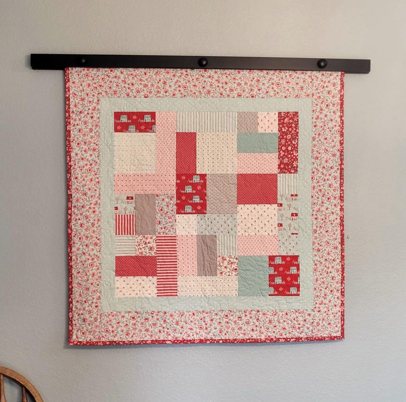 Peut inclure: Une courtepointe faite &agrave; la main avec un motif patchwork, compos&eacute;e de carr&eacute;s rouges, roses et gris. La courtepointe a une bordure florale et est suspendue &agrave; une tringle noire. Le motif comprend de petites maisons et des rayures.