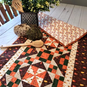May include: A quilted table runner featuring a patchwork pattern of orange, dark green, and brown squares with small house motifs. The runner is accented by two decorative ceramic pumpkins, a metal utensil holder with greenery, and a wooden spoon.