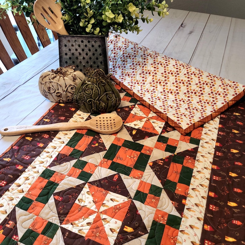 May include: A quilted table runner featuring a patchwork pattern of orange, dark green, and brown squares with small house motifs. The runner is accented by two decorative ceramic pumpkins, a metal utensil holder with greenery, and a wooden spoon.