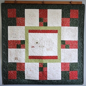May include: A Christmas-themed quilt with a dark green border and a patchwork design. The quilt features red and green squares, and white squares with embroidered Christmas scenes and text. The quilt is hung on a wooden rod.