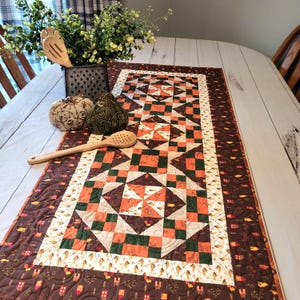 May include: A rectangular quilted table runner featuring a patchwork design in shades of orange, dark brown, and cream. The fabric pattern includes small house motifs and leaf prints, finished with intricate swirling machine quilting stitches.