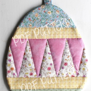 May include: A handmade quilted egg-shaped hot pad with a loop for hanging. The pad is made of various floral patterned fabrics in shades of blue, pink, yellow, and green.