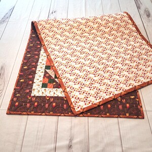 May include: A rectangular quilted table runner featuring a cream-colored top panel with a dense pattern of small red and yellow flowers. The border is dark brown with a repeating motif of tiny houses and trees, finished with a plaid fabric binding.