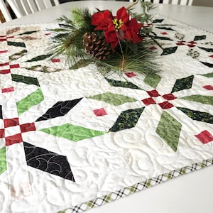 May include: A square quilted table topper featuring a white background with a repeating star pattern in shades of dark green, light green, black, and red. The fabric has intricate swirl stitching and a plaid border, topped with a pine and poinsettia sprig.