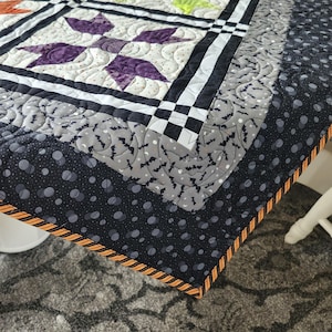 May include: A handmade quilt with a star pattern in the center, featuring purple and white floral designs. The border has a black and gray bat pattern, with a black and orange striped edge. The quilt is on a table.