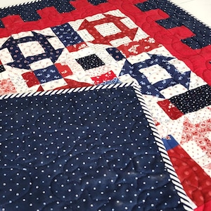 May include: A quilted blanket with a patriotic theme, featuring red, white, and blue patchwork. The design includes stars, squares, and geometric patterns. The reverse side is navy blue with white polka dots, and the edges are trimmed with a striped border.