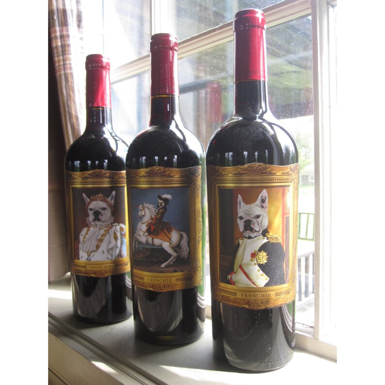 May include: Three bottles of red wine with gold labels featuring illustrations of French Bulldogs in various poses. The labels also include the text "Frenchie Wine".
