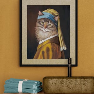 May include: Framed artwork featuring a cat in a yellow and blue head covering and attire, reminiscent of a classic painting. The cat has brown and white fur and is set against a dark background. The artwork is framed in a black frame.