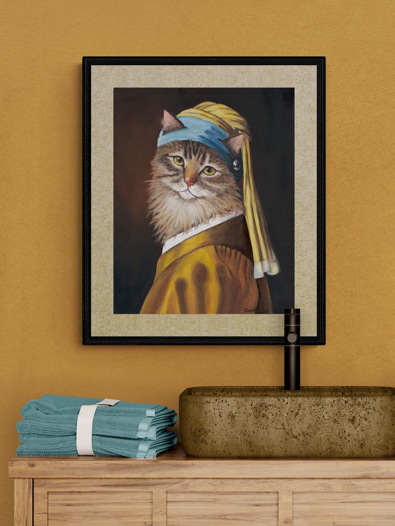 May include: Framed artwork featuring a cat in a yellow and blue head covering and attire, reminiscent of a classic painting. The cat has brown and white fur and is set against a dark background. The artwork is framed in a black frame.