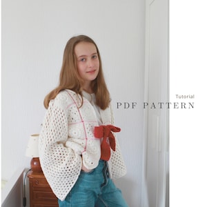 May include: A white crochet cardigan with a red and pink plaid pattern. The cardigan has a tie at the waist and bell sleeves. The model is wearing blue jeans.