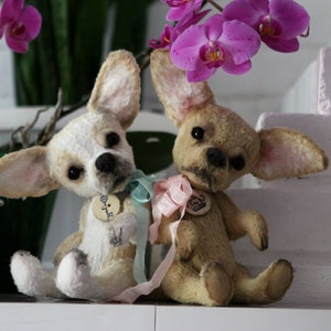 May include: Two plush chihuahua dog toys with light brown and white fur, wearing blue and pink ribbons. The toys are sitting on a white surface with a pink brick wall in the background.