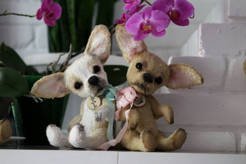 May include: Two plush chihuahua dog toys with light brown and white fur, wearing blue and pink ribbons. The toys are sitting on a white surface with a pink brick wall in the background.