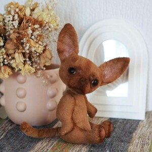 May include: A brown plush chihuahua dog toy with large ears and a sweet expression. The toy is sitting on a striped rug in front of a white mirror.