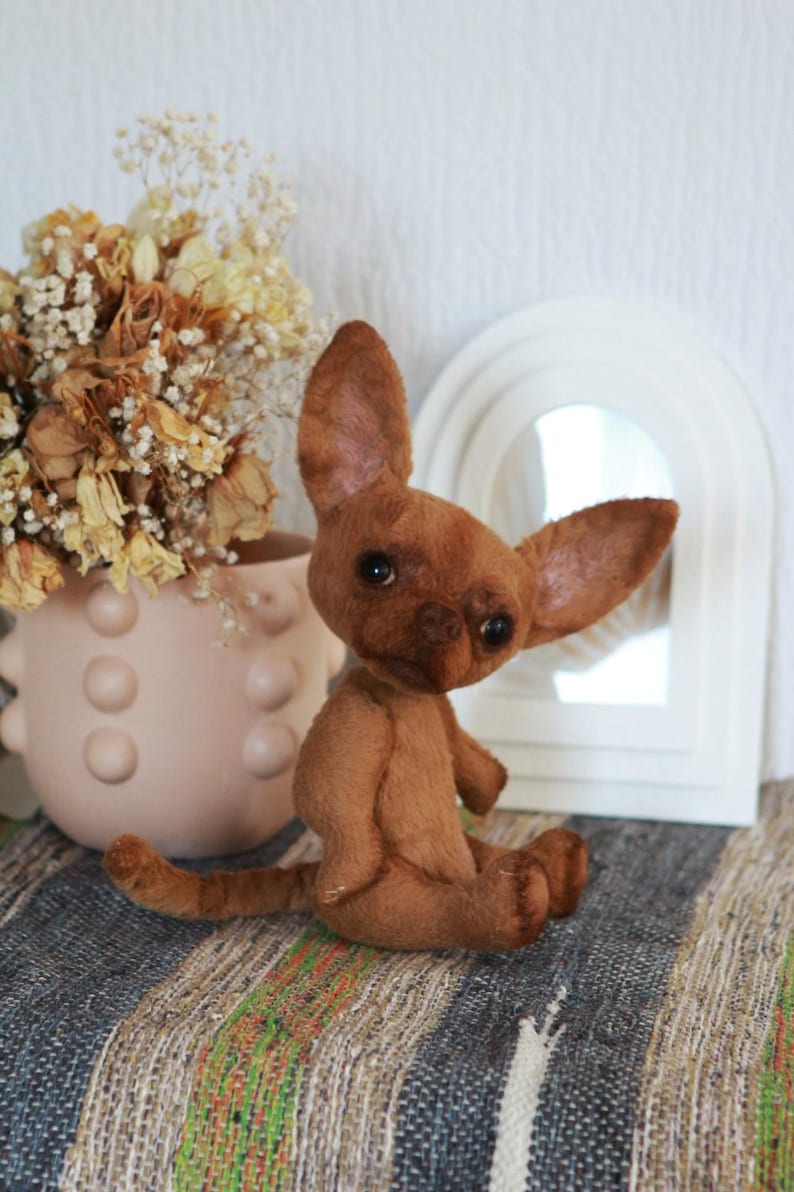 May include: A brown plush chihuahua dog toy with large ears and a sweet expression. The toy is sitting on a striped rug in front of a white mirror.