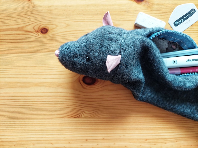 May include: A gray felt pencil case shaped like a mouse with pink ears and a black eye. The case is open and filled with pens and pencils. The case is sitting on a wooden surface.