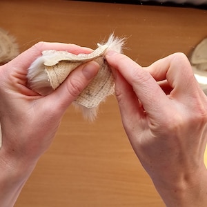 May include: A person is holding a white fur fabric with a beige backing. The fabric is folded in half and the person is holding the edges of the fabric.