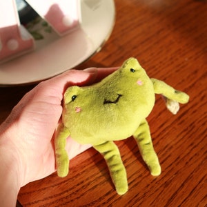 May include: A green plush frog toy with a smiling face and black eyes. The frog is being held in a hand.