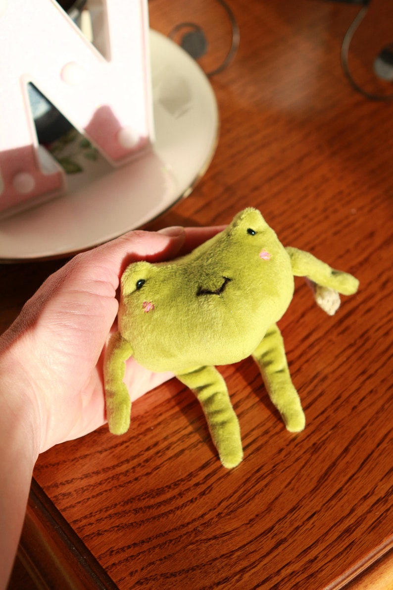 May include: A green plush frog toy with a smiling face and black eyes. The frog is being held in a hand.
