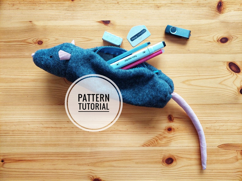 May include: A gray felt mouse-shaped pencil case with a zipper opening. The case is filled with pens, pencils, and other supplies. The text "PATTERN TUTORIAL" is written in a white circle on the case.