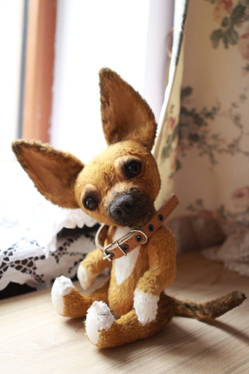 May include: A brown and white plush chihuahua dog toy with a brown leather collar. The toy is sitting on a wooden surface.