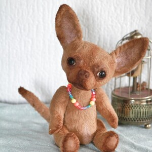 May include: A brown plush chihuahua toy with large eyes and a colorful beaded necklace. The toy is sitting on a light blue fabric surface.