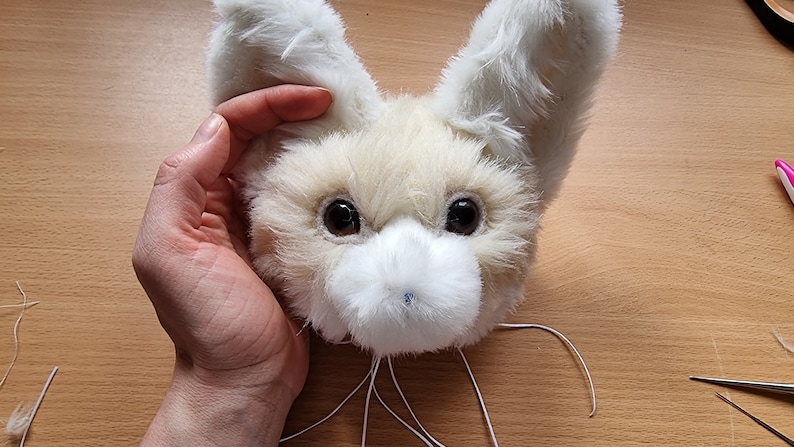 May include: A white fur head with large ears, brown eyes, and a white nose. The head is being held by a hand.
