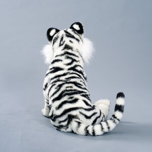 May include: A white and black plush tiger sitting on a gray background. The tiger is facing away from the camera, with its tail curled around its body.