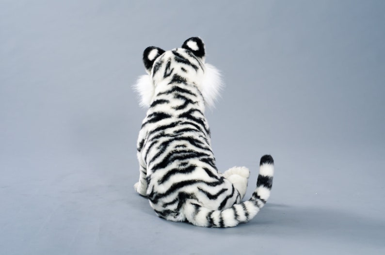 May include: A white and black plush tiger sitting on a gray background. The tiger is facing away from the camera, with its tail curled around its body.