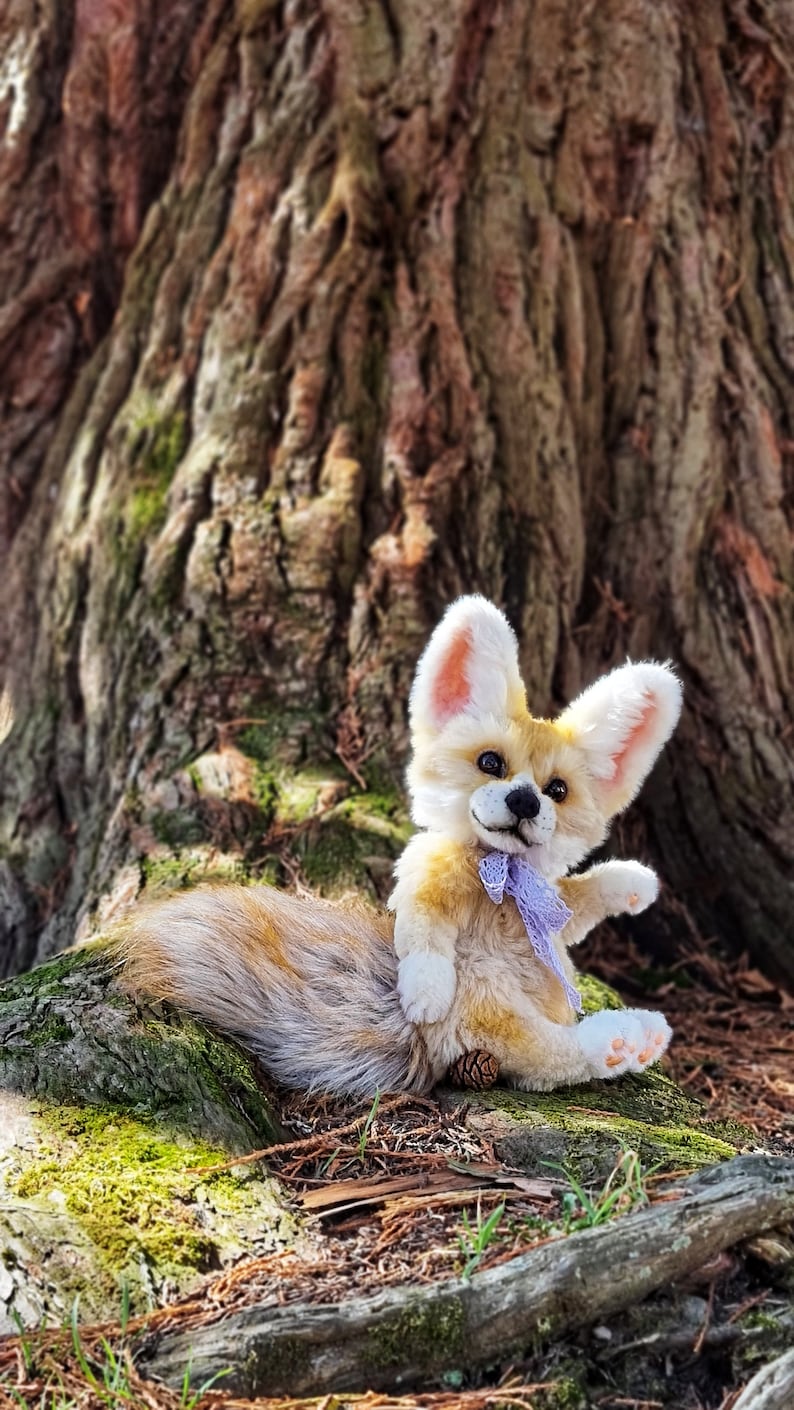 May include: A plush toy fox with a light brown and white coat, wearing a purple bow. The fox is sitting on a tree trunk with green moss and brown pine needles.