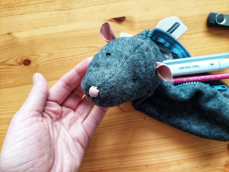 May include: A gray felt pencil case shaped like a mouse with pink ears and nose. The case is open and has several pens and pencils inside.