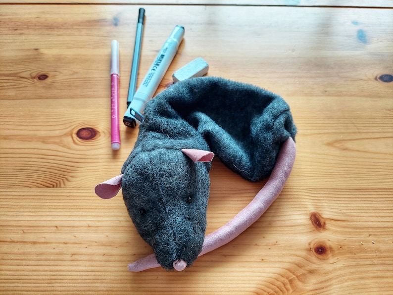 May include: A gray plush rat toy with a pink tail. The toy is lying on a wooden surface with three markers and a white eraser.