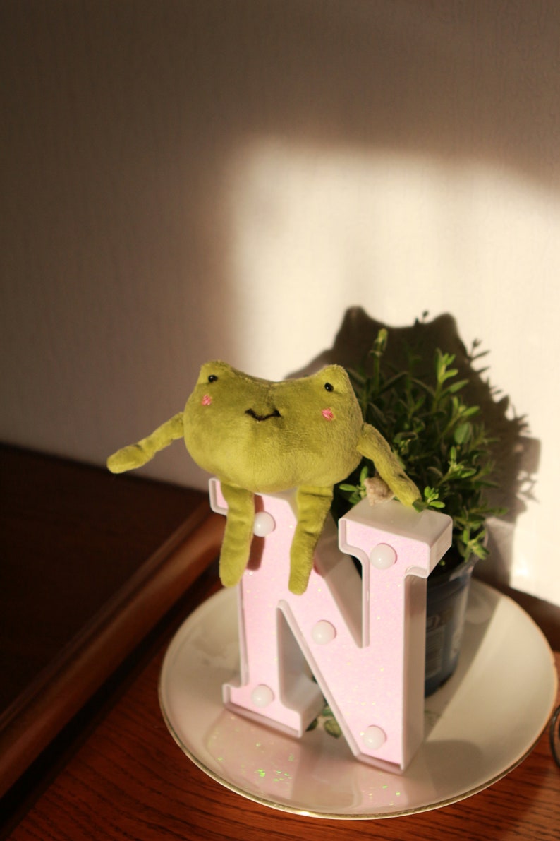 May include: A plush green frog toy sitting on a pink letter N with white lights. The letter is on a white saucer with a floral design.