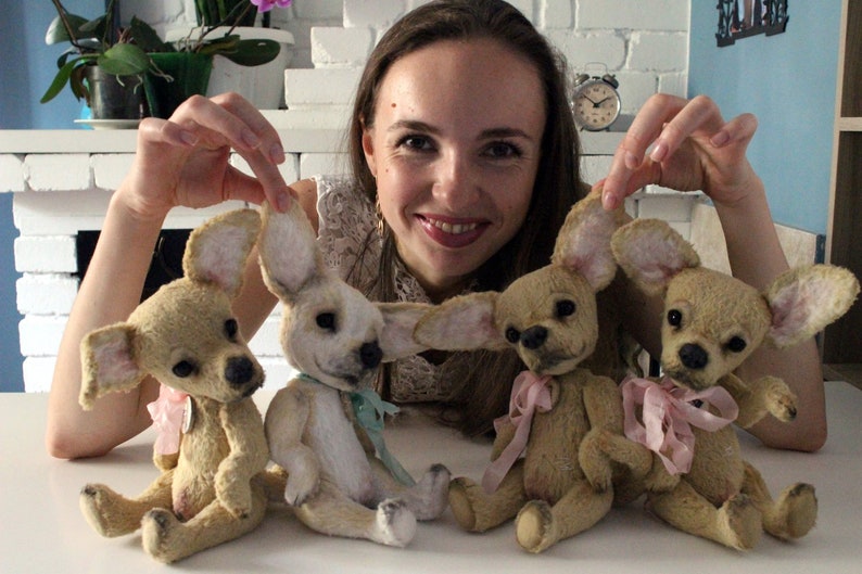 May include: Four handmade plush chihuahua dog toys. The toys are beige with pink and blue accents. The toys are sitting on a white surface.