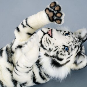 May include: A white tiger plush toy with black stripes, blue eyes, and a pink nose. The toy is lying on its back with its paws in the air.