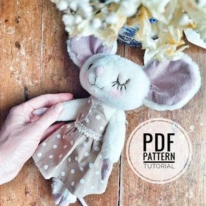 Op de afbeelding: Een handgemaakt wit pluche muisspeelgoed met grote, roze gevoerde oren, gekleed in een beige polka-dot jurk met kanten rand. De muis ligt op een houten oppervlak. De tekst "PDF PATTERN TUTORIAL" is inbegrepen.