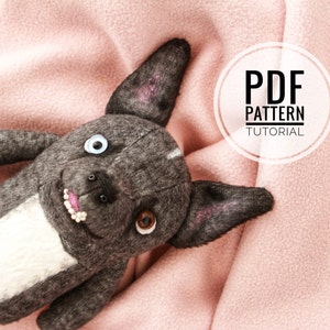 Puede incluir: Un perro de peluche gris con pelo blanco en el pecho, acostado sobre una manta de forro polar rosa. El perro tiene ojos grandes y redondos, uno azul y otro marrón, y una nariz rosa. La imagen incluye el texto "PDF PATTERN TUTORIAL" en un círculo blanco.