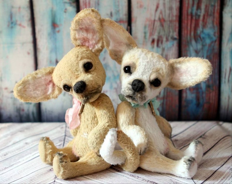 May include: Two plush chihuahua dog toys, one is cream colored with pink ears and a pink bow, the other is white with brown ears and a light blue bow.
