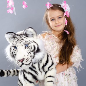 May include: A young girl wearing a white dress with floral print holds a white stuffed tiger with black stripes. She is wearing a headband with pink flowers. The background is a light gray with pink flower decorations hanging from the ceiling.