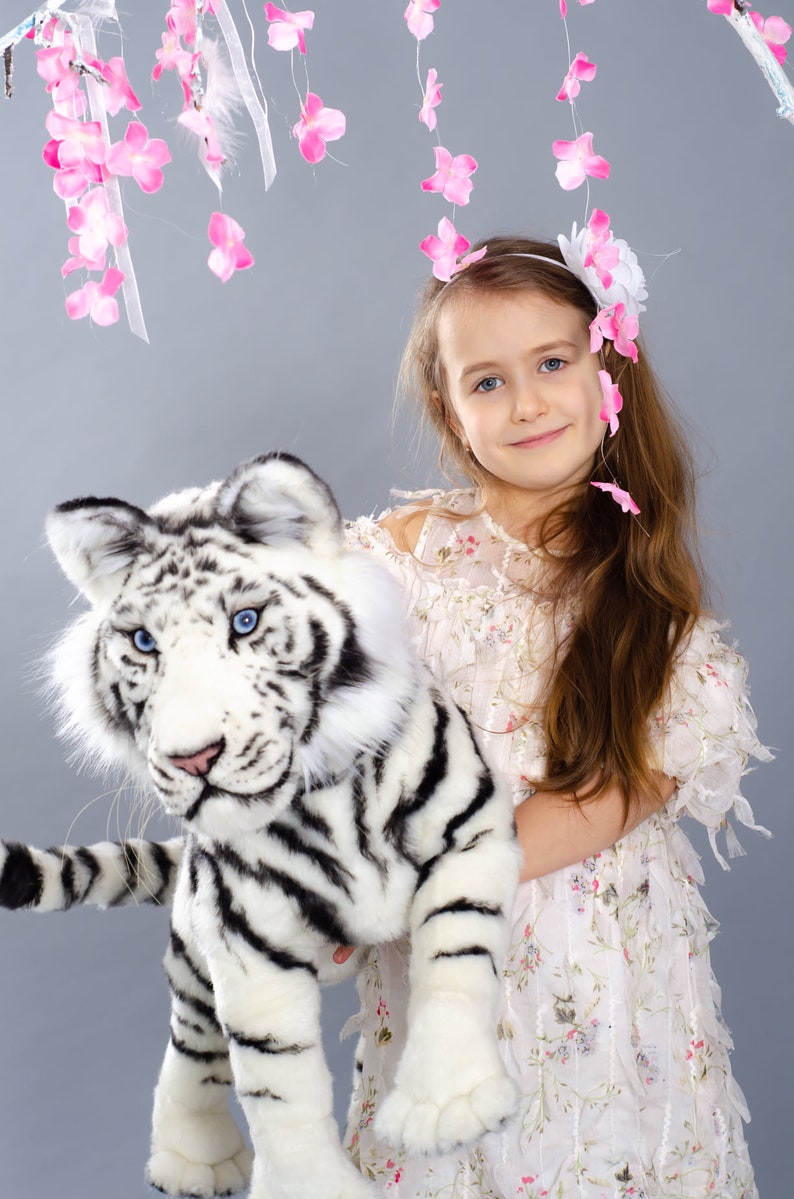 May include: A young girl wearing a white dress with floral print holds a white stuffed tiger with black stripes. She is wearing a headband with pink flowers. The background is a light gray with pink flower decorations hanging from the ceiling.