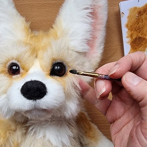 May include: A close-up of a plush toy fox with brown fur, white fur, and pink ears. The toy is being painted with a brown paintbrush. The paintbrush is being held by a hand.
